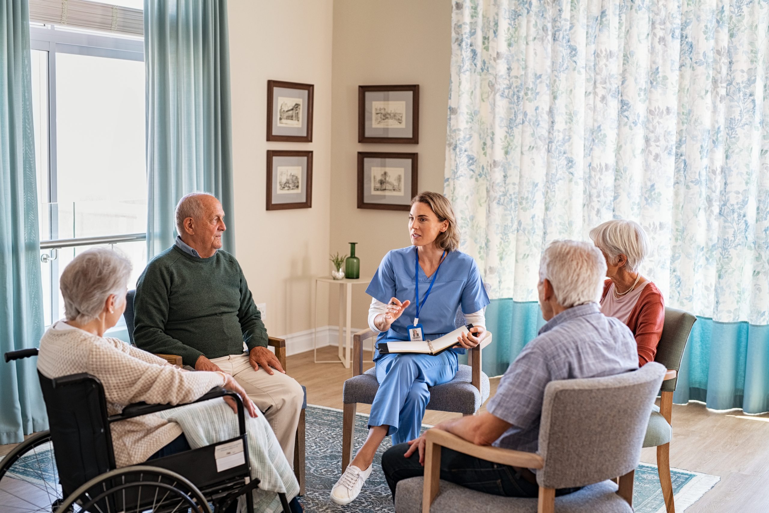 Group of senior people listening to young nurse. Psychological support group for elderly and lonely people in a community centre. Group therapy in session sitting in a circle in a nursing home facility.